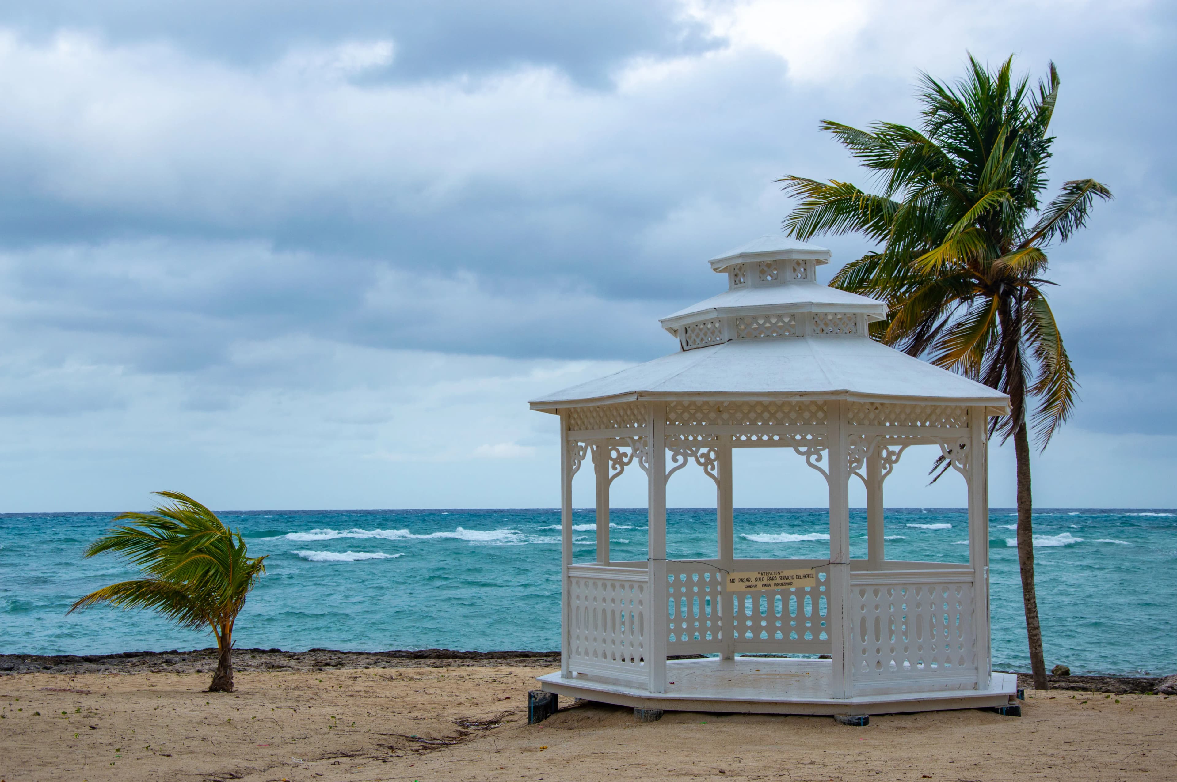 A sturdy, well-maintained Caribbean vacation rental home with hurricane shutters partially closed, surrounded by palm trees, under a calm sky.